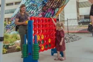 A young girl in a red dress plays a giant outdoor Connect Four game with a smiling woman nearby on the Encore Terrace at RBC Place London