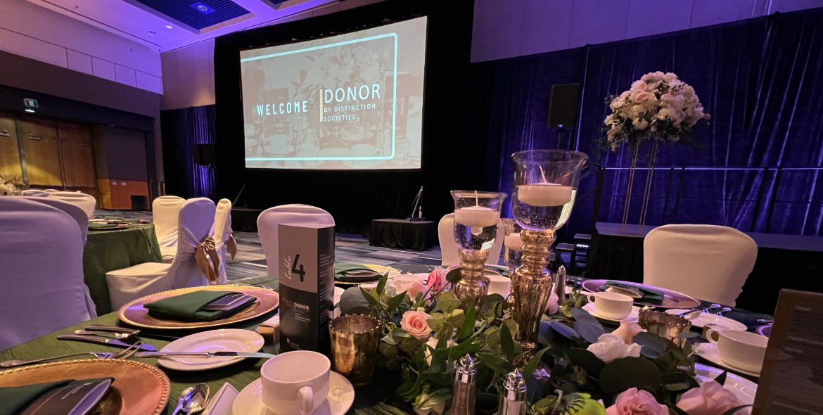 Elegant banquet table set with floral centerpieces and candles in a dimly lit ballroom featuring a large screen welcoming donors to the Donors of Distinction President's Dinner. This year's dinner will return to RBC Place London on May 13, 2026.